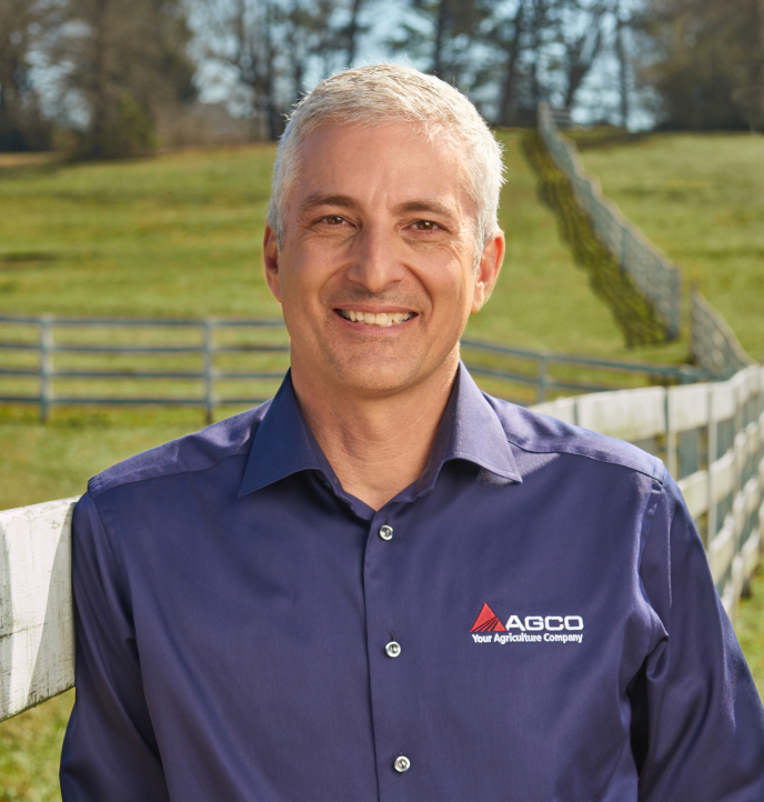Eric Hansotia穿着一件带有AGCO标志的蓝色衬衫，在绿色的田野里对着镜头微笑。